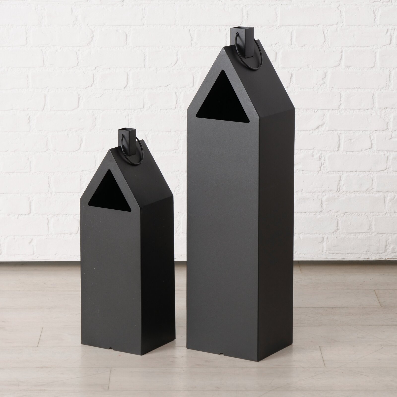 Twee zwarte, huisvormige metalen lantaarns van verschillende grootte met een driehoekige opening en een handvat, vrijstaand op een lichte ondergrond voor een witte bakstenen muur, vooraanzicht licht schuin van voren.