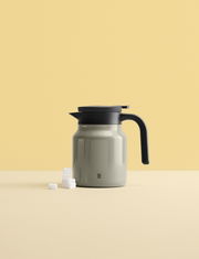 Graue Isolierkanne mit schwarzem Griff und Deckel, seitliche Ansicht, auf hellem Hintergrund mit Zuckerwürfeln.