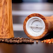 Pfeffermühle aus Olivenholz, 18 cm, liegend mit Blick auf das Keramikmahlwerk, umgeben von Pfefferkörnern.