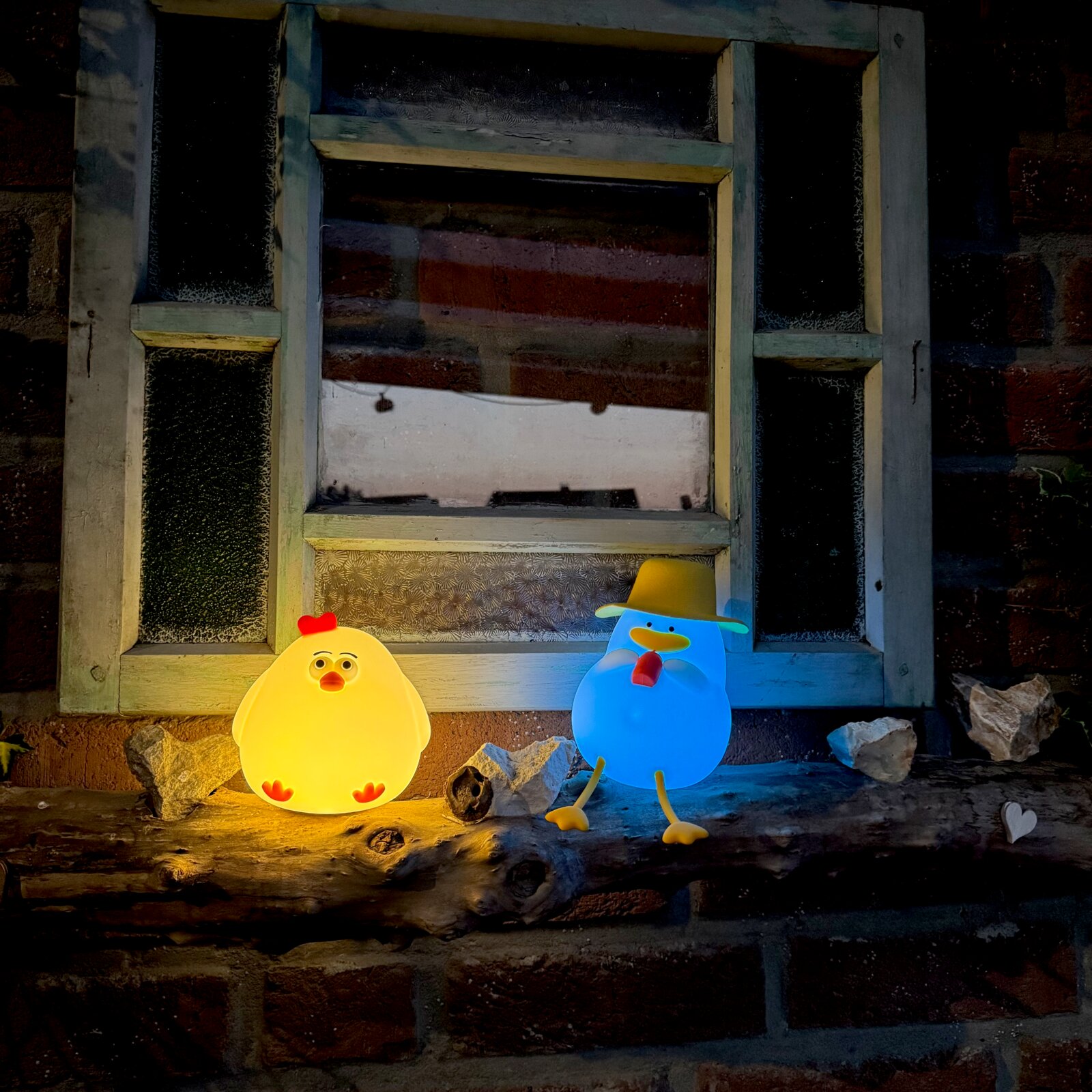 Zwei leuchtende Akku-Nachtlichter in Form von Enten, eine gelb und eine blau, auf einem Fensterbrett aus Holz, frontal betrachtet.