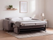 Grijze slaapbank met uitgeklapt metalen bedframe en wit beddengoed in een moderne woonkamer, gefotografeerd vanuit een licht zijdelings perspectief.