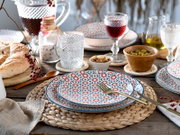 Rustikales Tischgedeck mit gemusterten Tellern in Blau-Rot, Glaswaren, Brot und Schalen mit Oliven und Nüssen auf Holztisch, aus leicht erhöhter schräger Perspektive