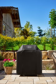 Abdeckhaube für BBQ-Station G4 in einem Garten, frontal fotografiert, mit einem Haus im Hintergrund.