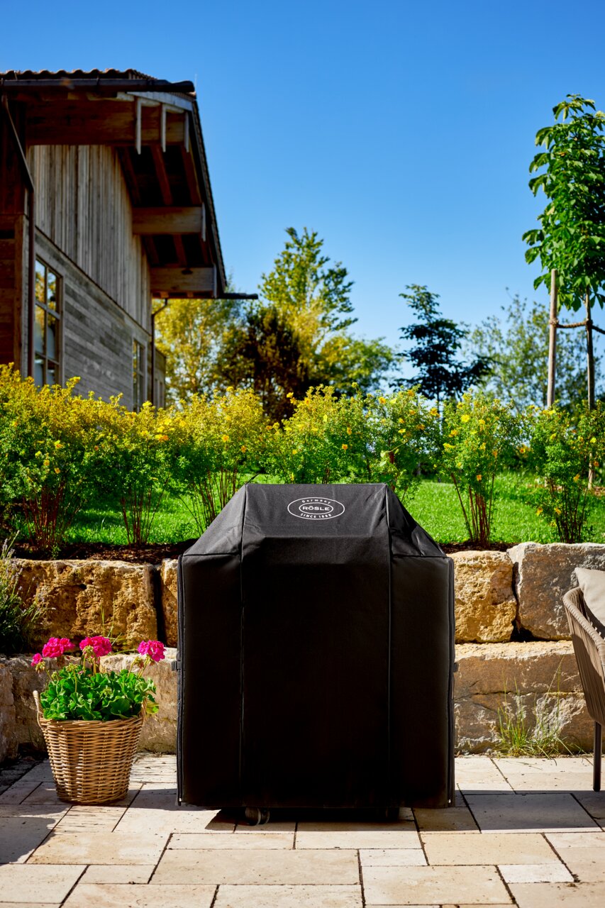 Rösle Abdeckhaube zu BBQ-Station VIDERO G4/G4-S & ALLFLAME 4 Abdeckhaube für BBQ-Station G4 in einem Garten, frontal fotografiert, mit einem Haus im Hintergrund.