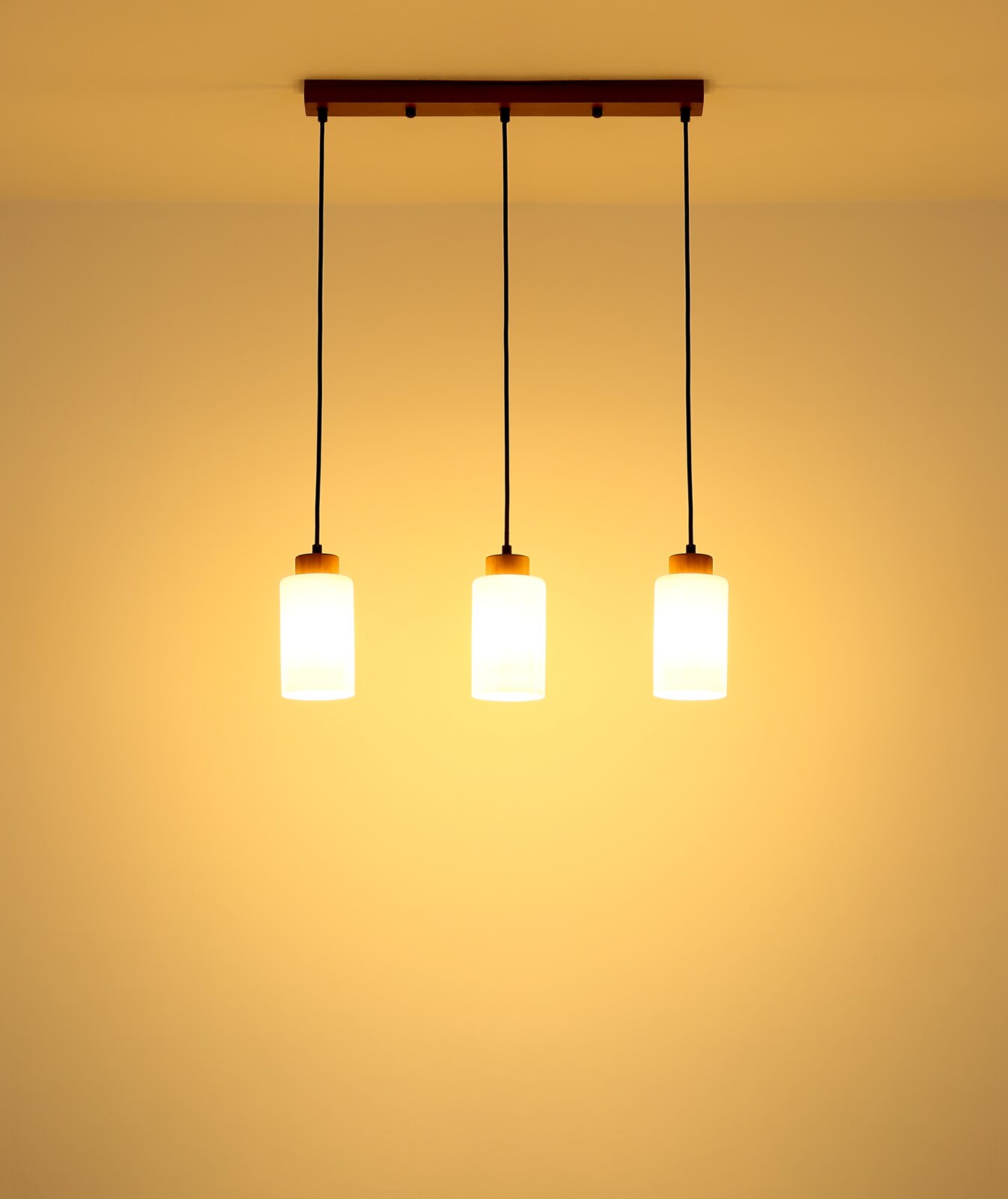 Hanglamp met drie lampen, voorzien van drie cilindervormige, opaalwitte glazen kappen aan een zwarte kabel en een rechthoekige houten kap, van voren en van onderaf tegen het plafond gefotografeerd, warm licht