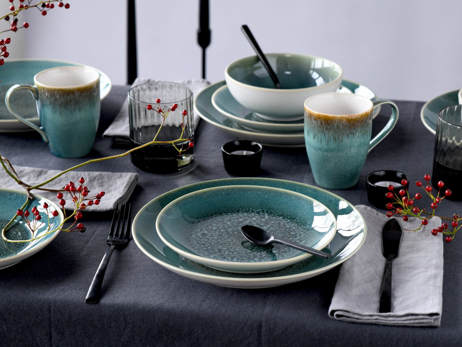 Schräg von vorn fotografierte Tischszene mit türkis-blau glasiertem Keramikgeschirr (Teller, Schalen, Tassen) auf dunkler Tischdecke, dazu schwarzes Besteck, geriffelte Gläser und rote Beerenzweige als Deko.