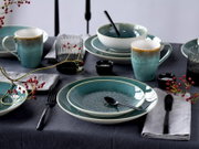 Schräg von vorn fotografierte Tischszene mit türkis-blau glasiertem Keramikgeschirr (Teller, Schalen, Tassen) auf dunkler Tischdecke, dazu schwarzes Besteck, geriffelte Gläser und rote Beerenzweige als Deko.