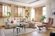 Gemütliches Wohnzimmer mit einer großen, beigen Rundecke, dekoriert mit Kissen und einer Decke. Im Vordergrund sind zwei schwarze Couchtische und ein gemusterter Teppich zu sehen. Die Perspektive zeigt den Raum von vorne, mit Blick auf die Fenster mit Hol
