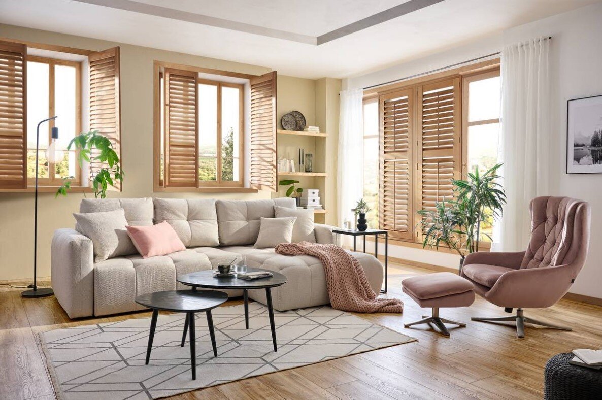 Gemütliches Wohnzimmer mit einer großen, beigen Rundecke, dekoriert mit Kissen und einer Decke. Im Vordergrund sind zwei schwarze Couchtische und ein gemusterter Teppich zu sehen. Die Perspektive zeigt den Raum von vorne, mit Blick auf die Fenster mit Hol