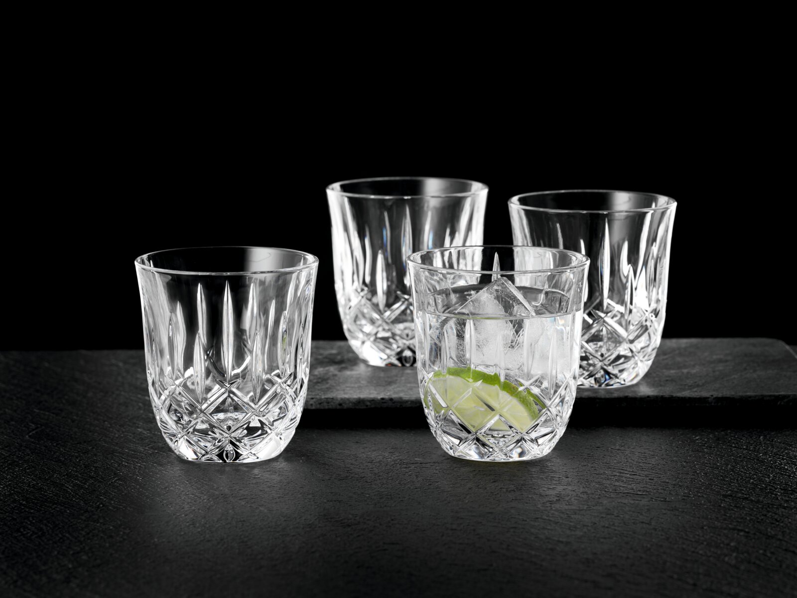 Vier klare Kristall-Whiskygläser mit Facettenschliff auf dunkler Steinplatte, eines mit Eiswürfeln und Limettenscheibe frontale Produktansicht leicht von oben