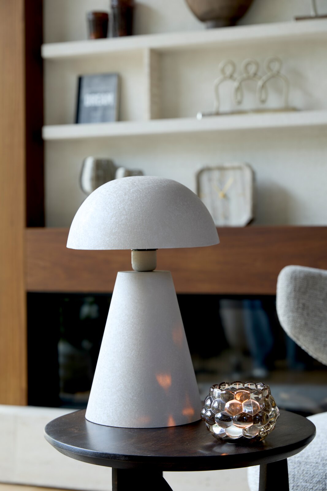 Light&Living Tafellamp SURRINA Moderne tafellamp in kegelvorm met een ronde kap, gezien vanaf de zijkant, geplaatst op een donkere houten tafel, met op de achtergrond een plank met decoratie.