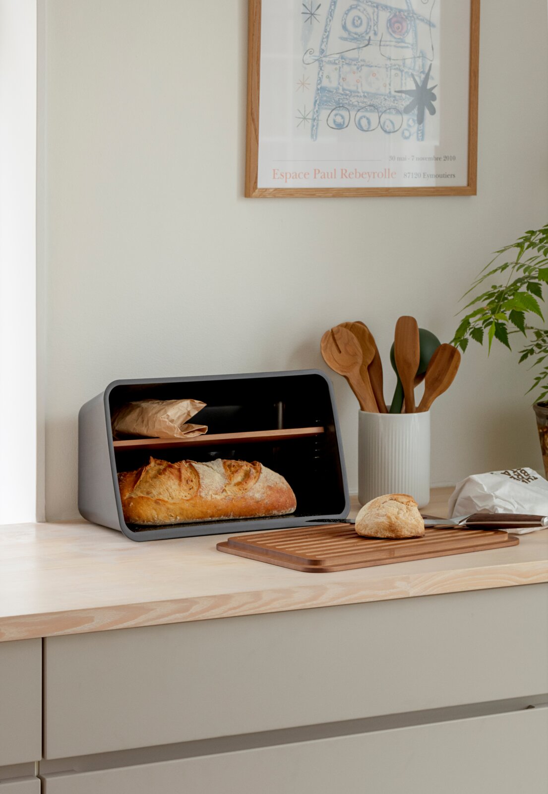 Moderner Brotkasten aus grauem Metall mit Holzgriff, seitliche Perspektive auf der Küchenarbeitsplatte innen liegt ein Laib Brot, daneben Schneidebrett mit Brötchen und Messer