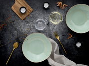 Draufsicht auf ein stilvolles Tisch-Arrangement mit zwei mintgrünen Keramikschalen, goldfarbenem Besteck, Leinenserviette, Wasserglas, Teelichtern und Holzbrett auf dunkler Oberfläche.
