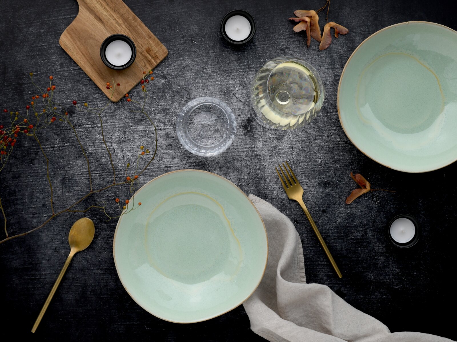 Draufsicht auf ein stilvolles Tisch-Arrangement mit zwei mintgrünen Keramikschalen, goldfarbenem Besteck, Leinenserviette, Wasserglas, Teelichtern und Holzbrett auf dunkler Oberfläche.