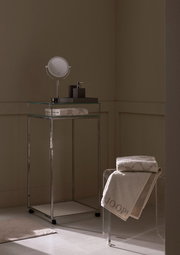 Luxuriöses Gästetuch in einem stilvollen Badezimmer, auf einem transparenten Stuhl drapiert, mit einem modernen Waschtisch im Hintergrund. Perspektive von der Seite.