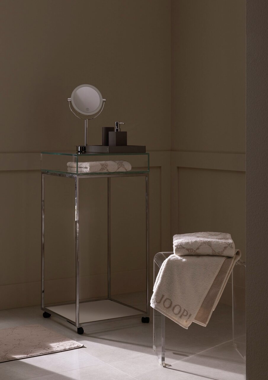 Luxuriöses Gästetuch in einem stilvollen Badezimmer, auf einem transparenten Stuhl drapiert, mit einem modernen Waschtisch im Hintergrund. Perspektive von der Seite.