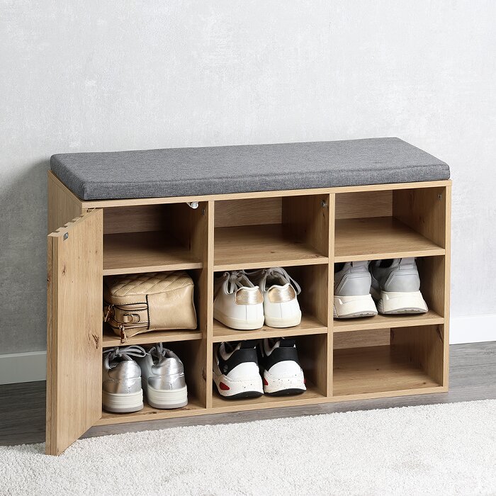 Houten schoenenbank met grijs zitkussen en open opbergvakken, linkerdeur open met schoenen en tas erin, gefotografeerd schuin van voren op ooghoogte tegen een lichtgrijze muur.
