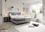 Modernes Boxspringbett in einem hellen Schlafzimmer, seitliche Perspektive, mit grauem Kopfteil und verstellbaren Matratzen, umgeben von stilvoller Dekoration.