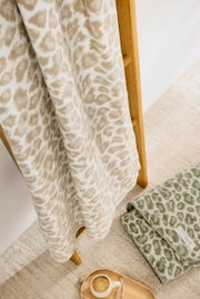 Leopardengemusterte Wohndecke in Beige auf Holzleiter, daneben eine grüne Decke und ein Tablett mit Kaffee, aus der Vogelperspektive.