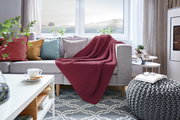 Gemütliches Wohnzimmer mit einer roten Strickdecke auf einem grauen Sofa, umgeben von bunten Kissen. Die Perspektive zeigt die Decke von vorne, drapiert über die Sofalehne.