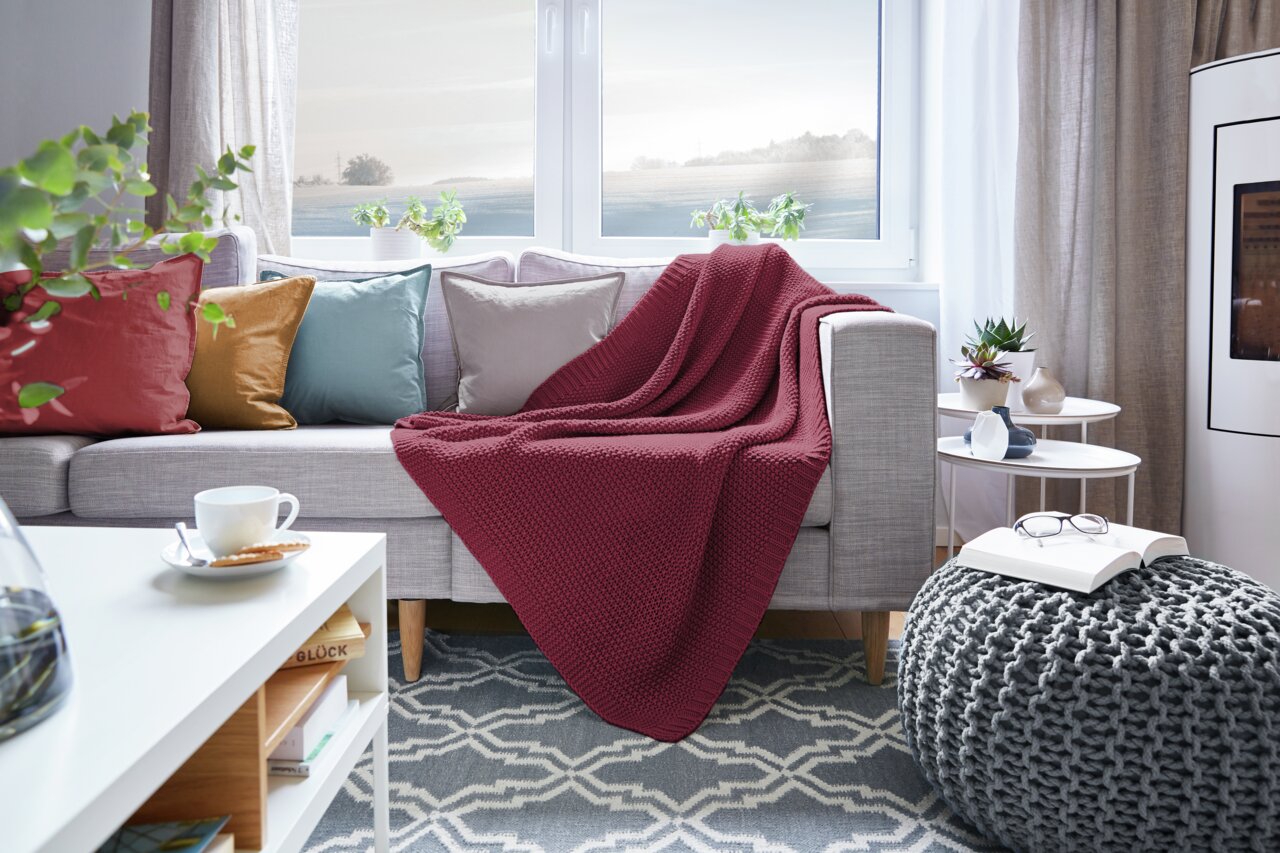 Gemütliches Wohnzimmer mit einer roten Strickdecke auf einem grauen Sofa, umgeben von bunten Kissen. Die Perspektive zeigt die Decke von vorne, drapiert über die Sofalehne.