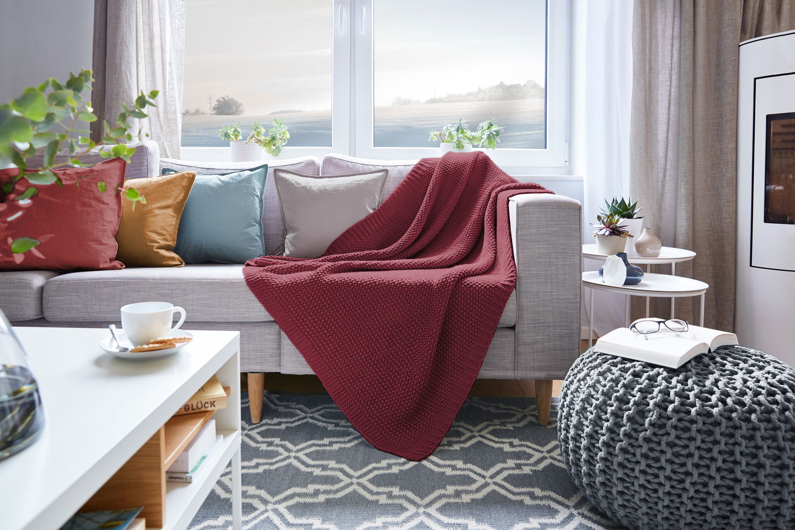 Gemütliches Wohnzimmer mit einer roten Strickdecke auf einem grauen Sofa, umgeben von bunten Kissen. Die Perspektive zeigt die Decke von vorne, drapiert über die Sofalehne.