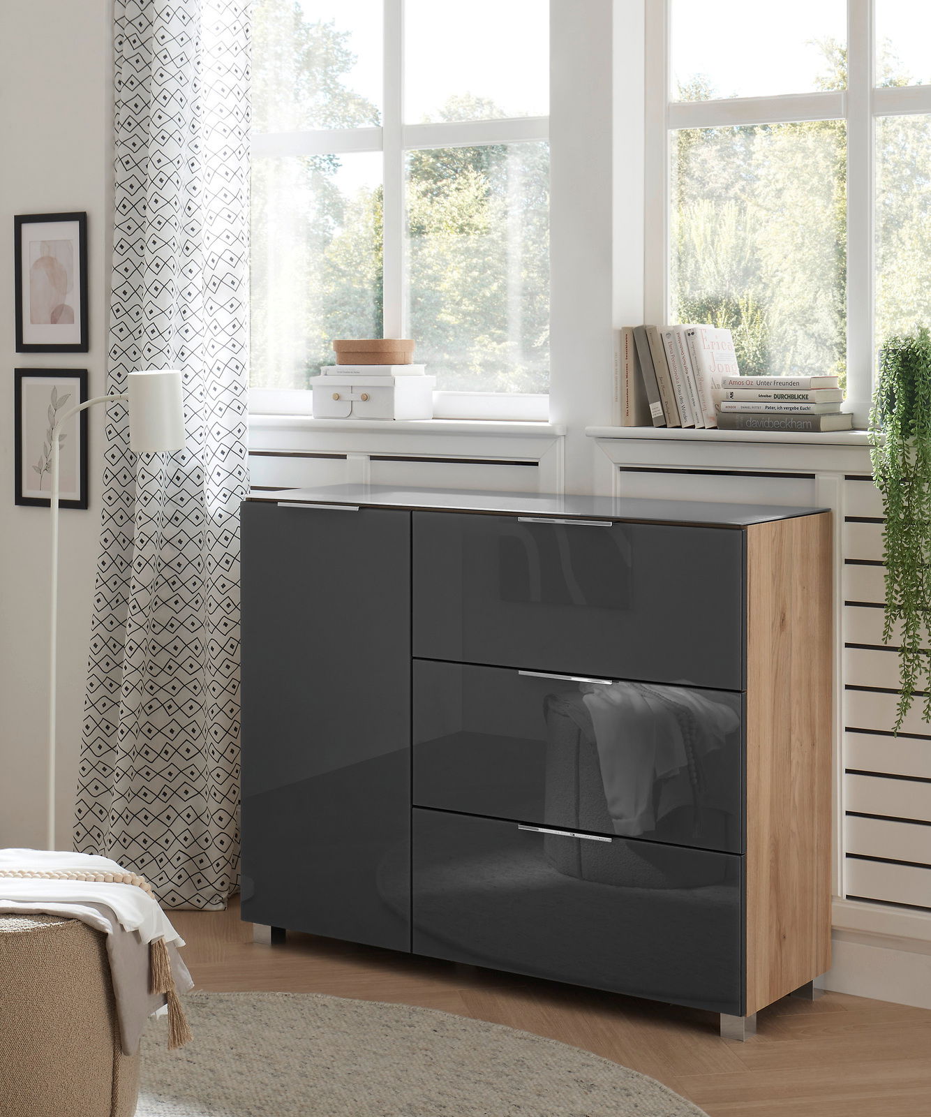 Moderne Kommode mit glänzenden, dunklen Fronten und Holzseiten, in einem hellen Raum mit großen Fenstern, aus seitlicher Perspektive fotografiert.