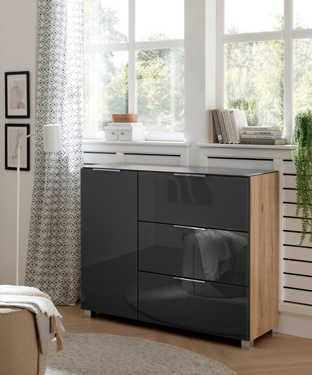 Moderne Kommode mit glänzenden, dunklen Fronten und Holzseiten, in einem hellen Raum mit großen Fenstern, aus seitlicher Perspektive fotografiert.