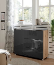 Moderne Kommode mit glänzenden, dunklen Fronten und Holzseiten, in einem hellen Raum mit großen Fenstern, aus seitlicher Perspektive fotografiert.