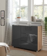 Moderne Kommode mit glänzenden, dunklen Fronten und Holzseiten, in einem hellen Raum mit großen Fenstern, aus seitlicher Perspektive fotografiert.