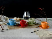 Vier bunte Espresso-/Kaffeetassen mit Untertassen (blau, rot, grün, orange) auf Holztablett auf gedecktem Tisch, mit Gläsern und Gebäck im Hintergrund; Perspektive: leicht erhöhte Frontansicht.