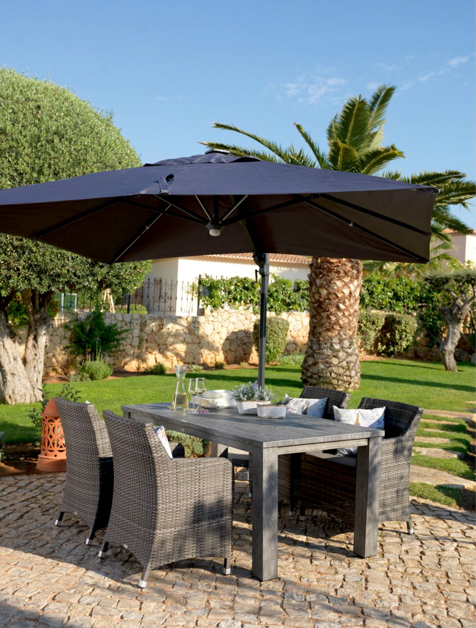 Großer Ampelschirm über einem Gartentisch mit vier geflochtenen Stühlen, umgeben von mediterraner Gartenlandschaft, seitliche Perspektive.