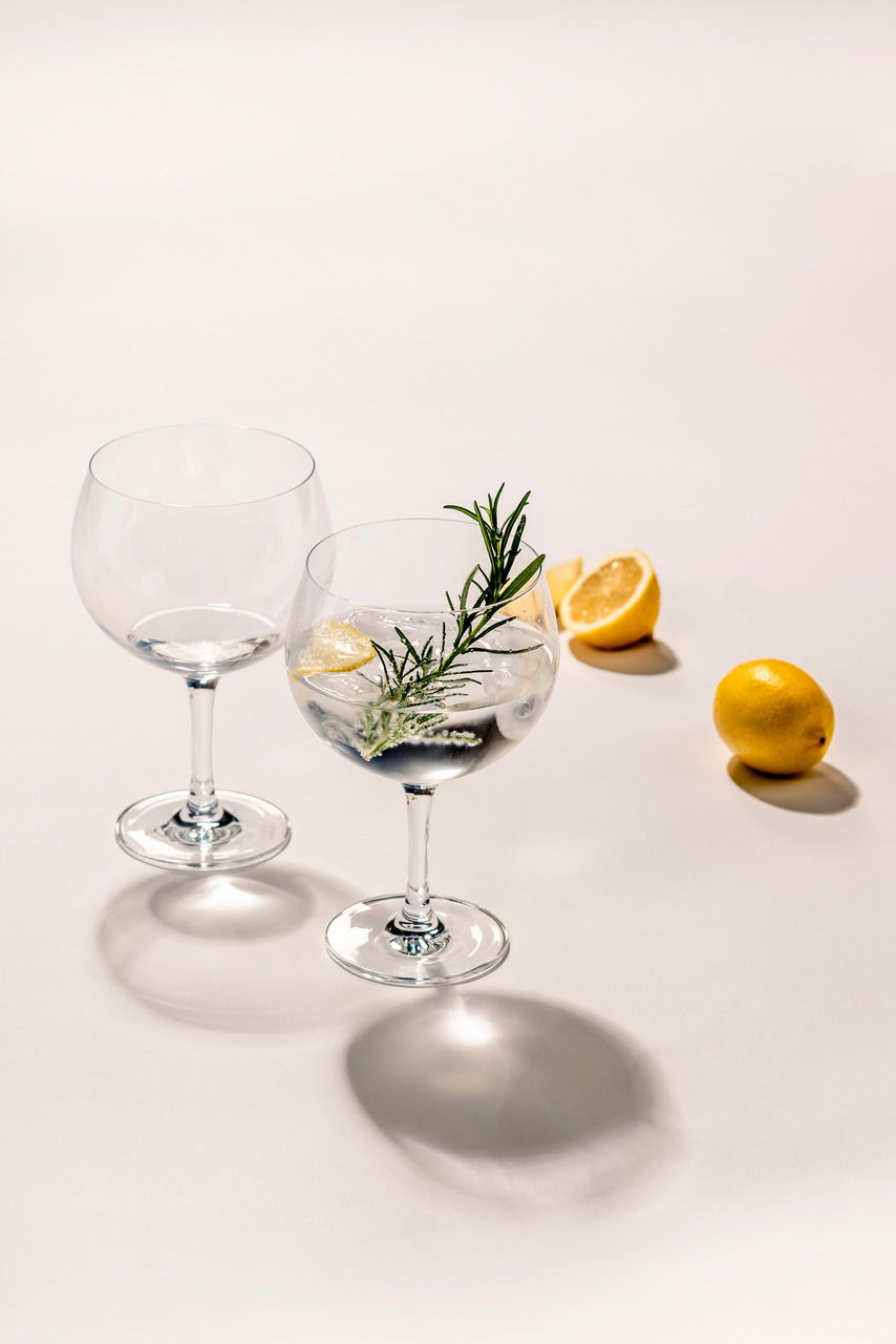 Zwei elegante Weißweingläser, eines gefüllt mit einem Getränk, Eiswürfeln, Rosmarin und Zitronenscheiben, auf einer hellen Oberfläche mit Zitronen im Hintergrund. Seitliche Perspektive.