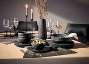 Elegantes 16-teiliges Geschirrset 'Manufacture Rock' in moderner Steinoptik, präsentiert auf einem stilvoll gedeckten Tisch mit Kerzen und Weingläsern, aus seitlicher Perspektive.
