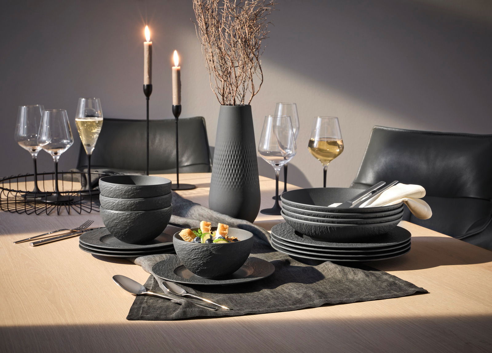 Elegantes 16-teiliges Geschirrset 'Manufacture Rock' in moderner Steinoptik, präsentiert auf einem stilvoll gedeckten Tisch mit Kerzen und Weingläsern, aus seitlicher Perspektive.