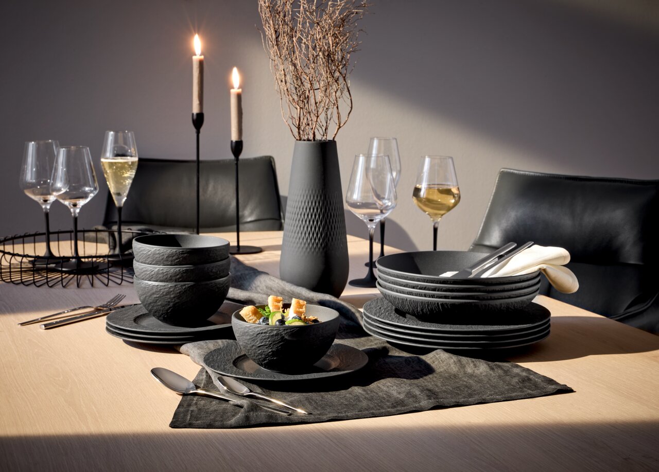 Elegante 16-delige servieset 'Manufacture Rock' in een moderne steenlook, gepresenteerd op een stijlvol gedekte tafel met kaarsen en wijnglazen, gezien vanuit een zijdelingse hoek.