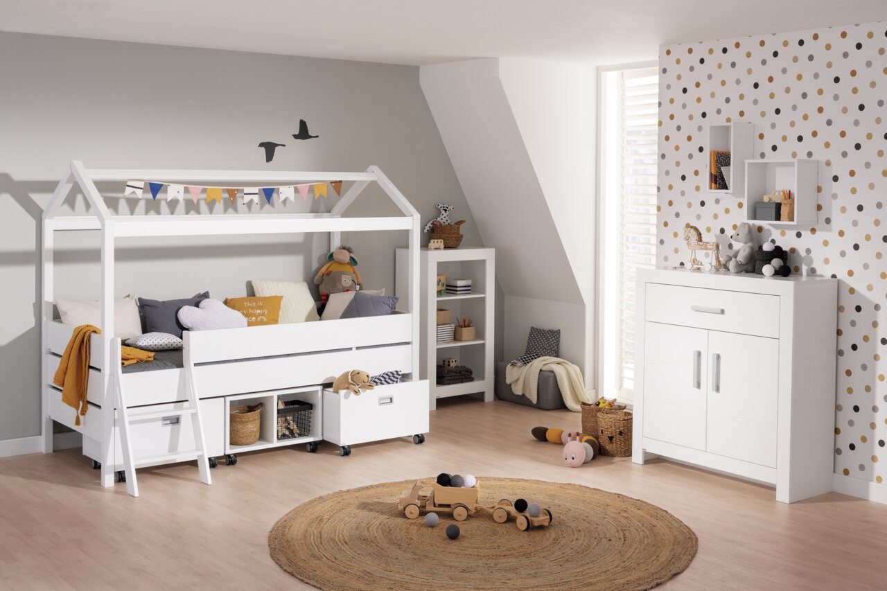 Paidi Stapelbed met frontpaneel TINY-HOUSE Kojenbett mit Giebel in einem Kinderzimmer, seitliche Perspektive. Das Bett ist weiß und mit bunten Wimpeln dekoriert. Darunter befinden sich Schubladen und Körbe zur Aufbewahrung. Im Hintergrund sind ein Regal und eine Kommode zu sehen.