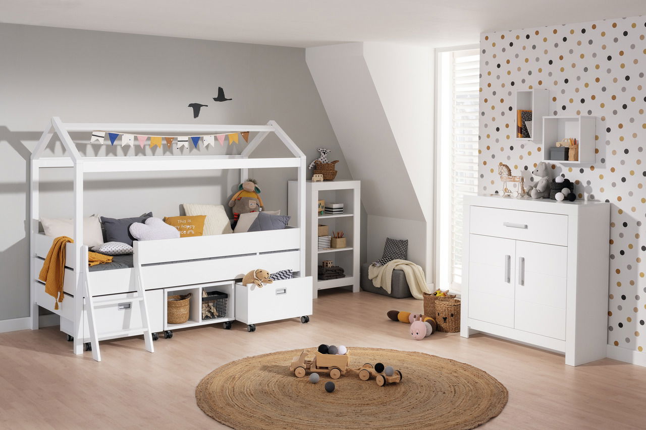 Kojenbett mit Giebel in einem Kinderzimmer, seitliche Perspektive. Das Bett ist weiß und mit bunten Wimpeln dekoriert. Darunter befinden sich Schubladen und Körbe zur Aufbewahrung. Im Hintergrund sind ein Regal und eine Kommode zu sehen.