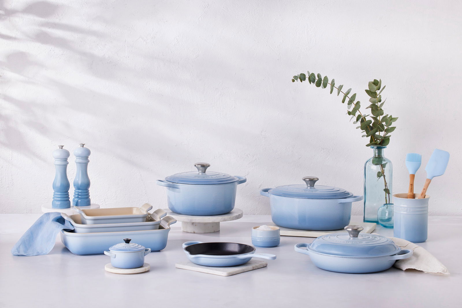 Eine Kollektion von hellblauen Küchenutensilien, darunter Bräter, Töpfe und Pfannen, auf einer weißen Oberfläche, seitlich von vorne fotografiert.