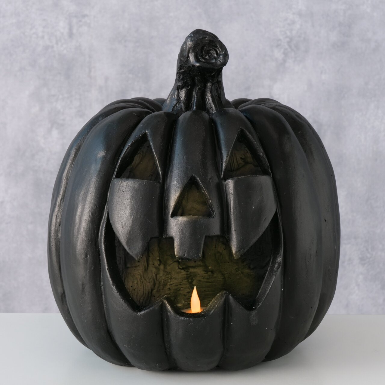Schwarzer Halloween-Kürbis (Jack-o’-Lantern) als Deko mit ausgeschnittenem Gesicht und kleiner Kerzenflamme im Inneren, frontal aus leicht erhöhter Perspektive vor grauem Hintergrund