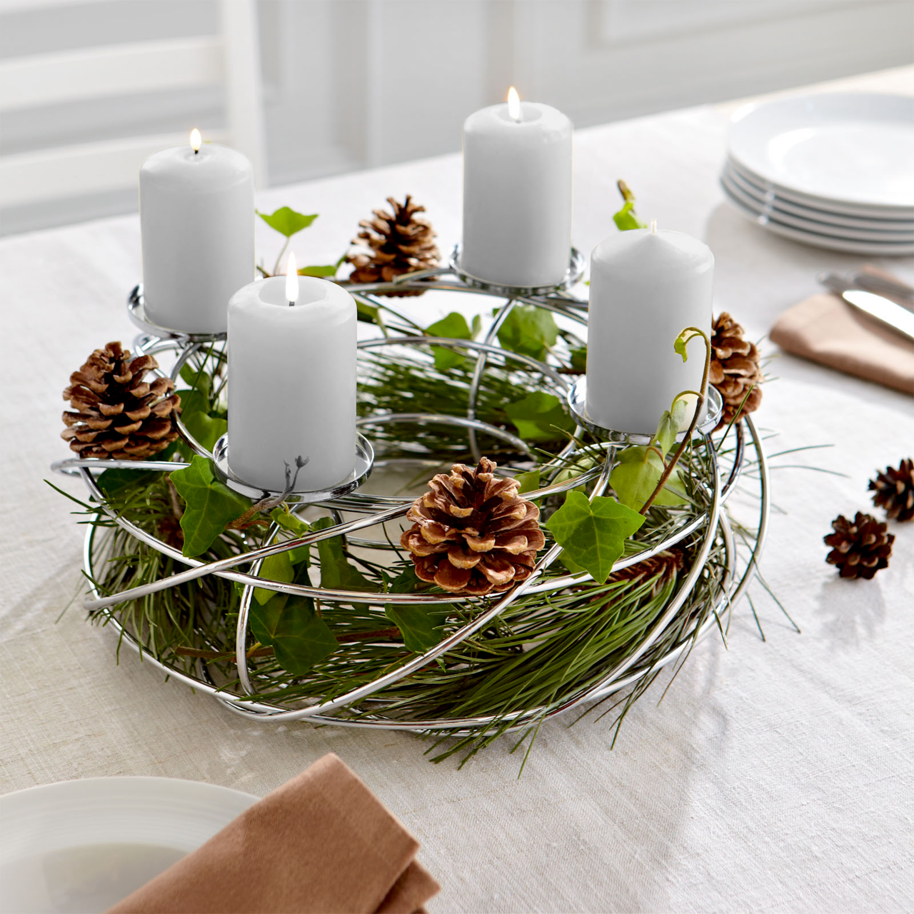 Silberner Dekokranz für 4 Kerzen mit Tannenzapfen und grünen Zweigen, auf einem Tisch arrangiert. Perspektive von schräg oben.