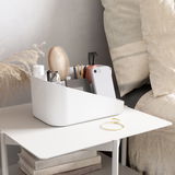 Weißer Organizer für Haarwerkzeuge auf einem Tisch neben einem Bett, gefüllt mit Bürsten, Flaschen und Accessoires, aus seitlicher Perspektive.
