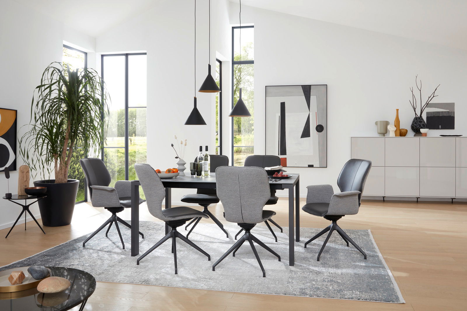 Moderner Essbereich mit sechs grauen Armlehnstühlen um einen schwarzen Tisch, helle und luftige Atmosphäre mit großen Fenstern im Hintergrund, seitliche Perspektive.