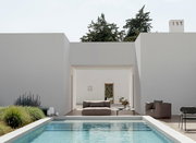 Frontalansicht auf einen modernen, weiß verputzten Innenhof mit rechteckigem Pool im Vordergrund, Loungesofa und Sessel sowie Beistelltisch auf der Terrasse, im Hintergrund Bäume und ein Eingang.