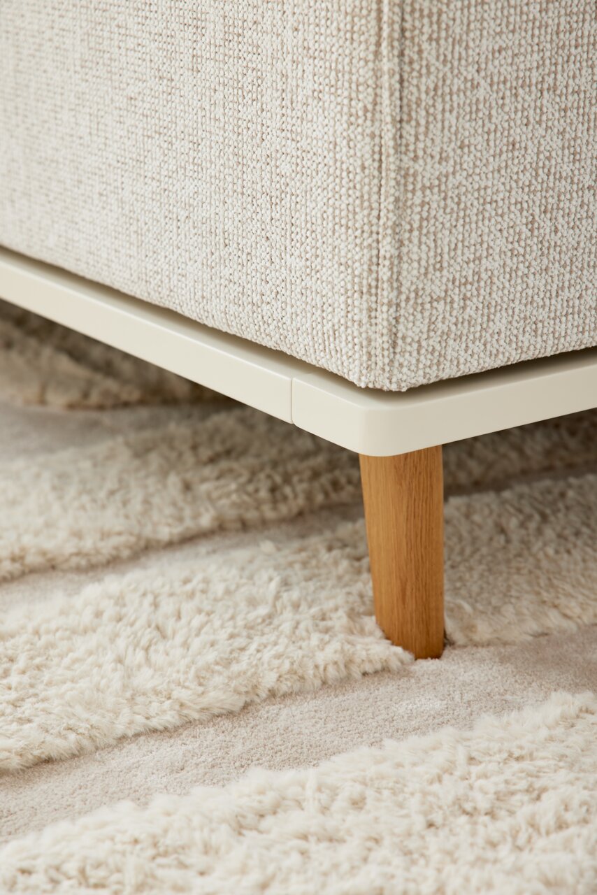 Close-up van een gestoffeerd bed met een lichtbeige stoffen bekleding en een houten onderstel, gezien vanaf de zijkant.