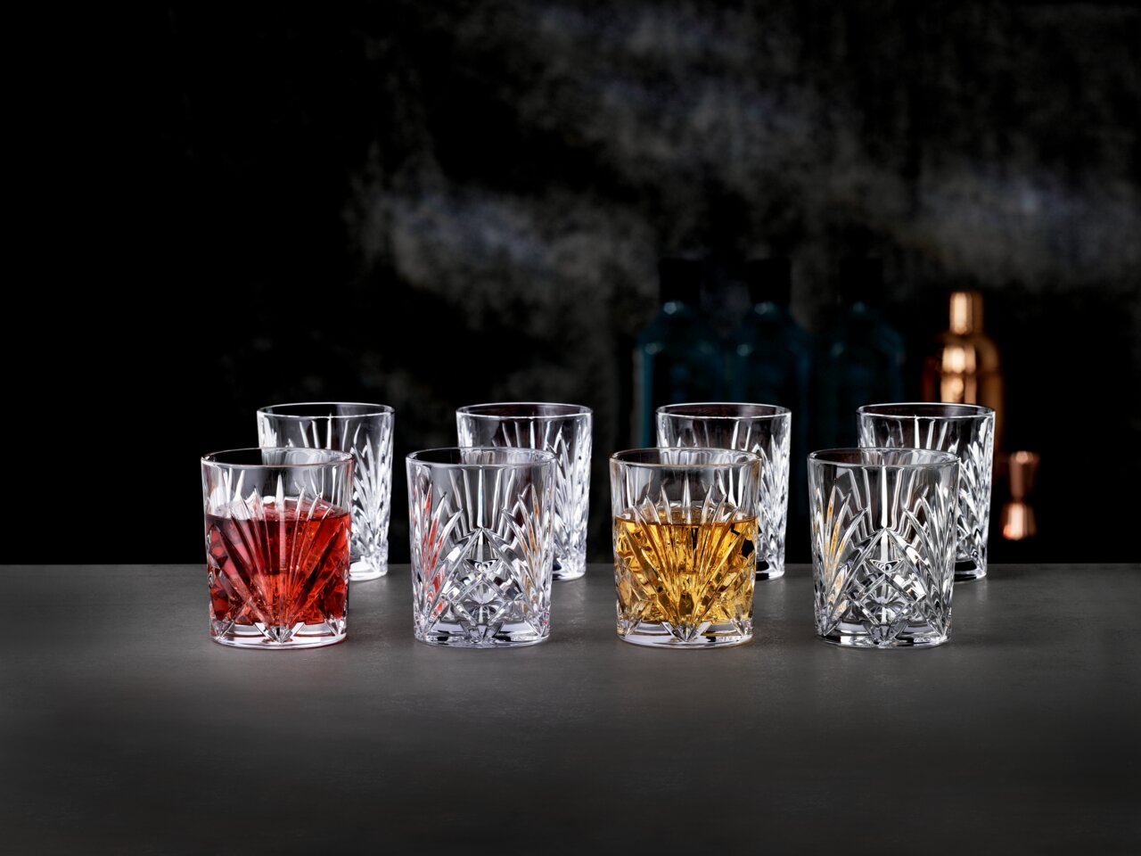 Sechs kristallklare Whisky-Tumbler mit geschliffenem Facettenschliff auf dunkler Oberfläche, zwei Gläser mit rotem und bernsteinfarbenem Drink, fotografiert in frontaler Perspektive auf Augenhöhe.