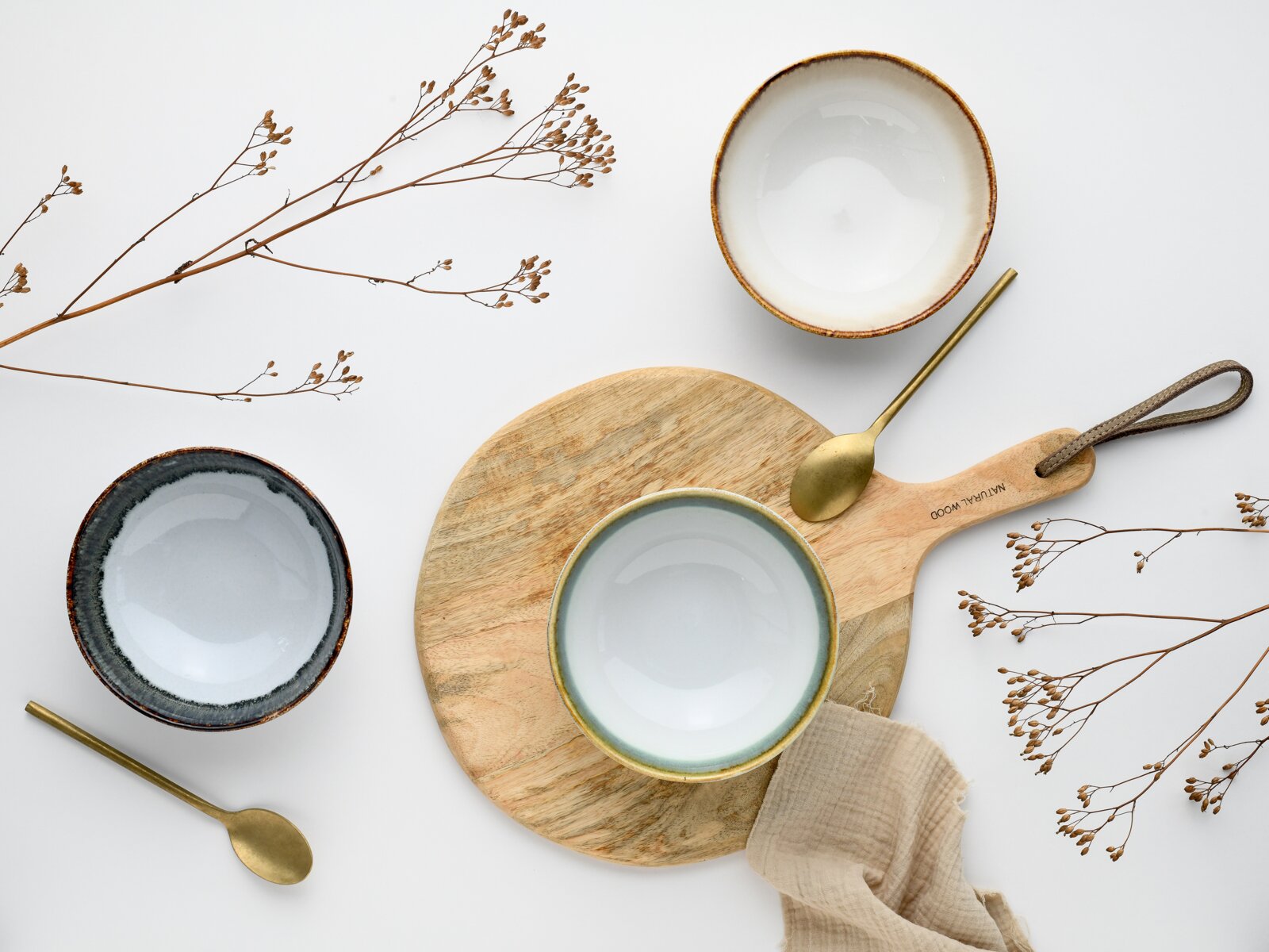 Drei Keramikschalen in Weiß und Grau mit braunem Rand, goldfarbene Löffel und Holz-Servierbrett mit Stofftuch und Trockenblumen, Draufsicht (Flatlay) auf hellem Hintergrund.
