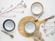 Drei Keramikschalen in Weiß und Grau mit braunem Rand, goldfarbene Löffel und Holz-Servierbrett mit Stofftuch und Trockenblumen, Draufsicht (Flatlay) auf hellem Hintergrund.