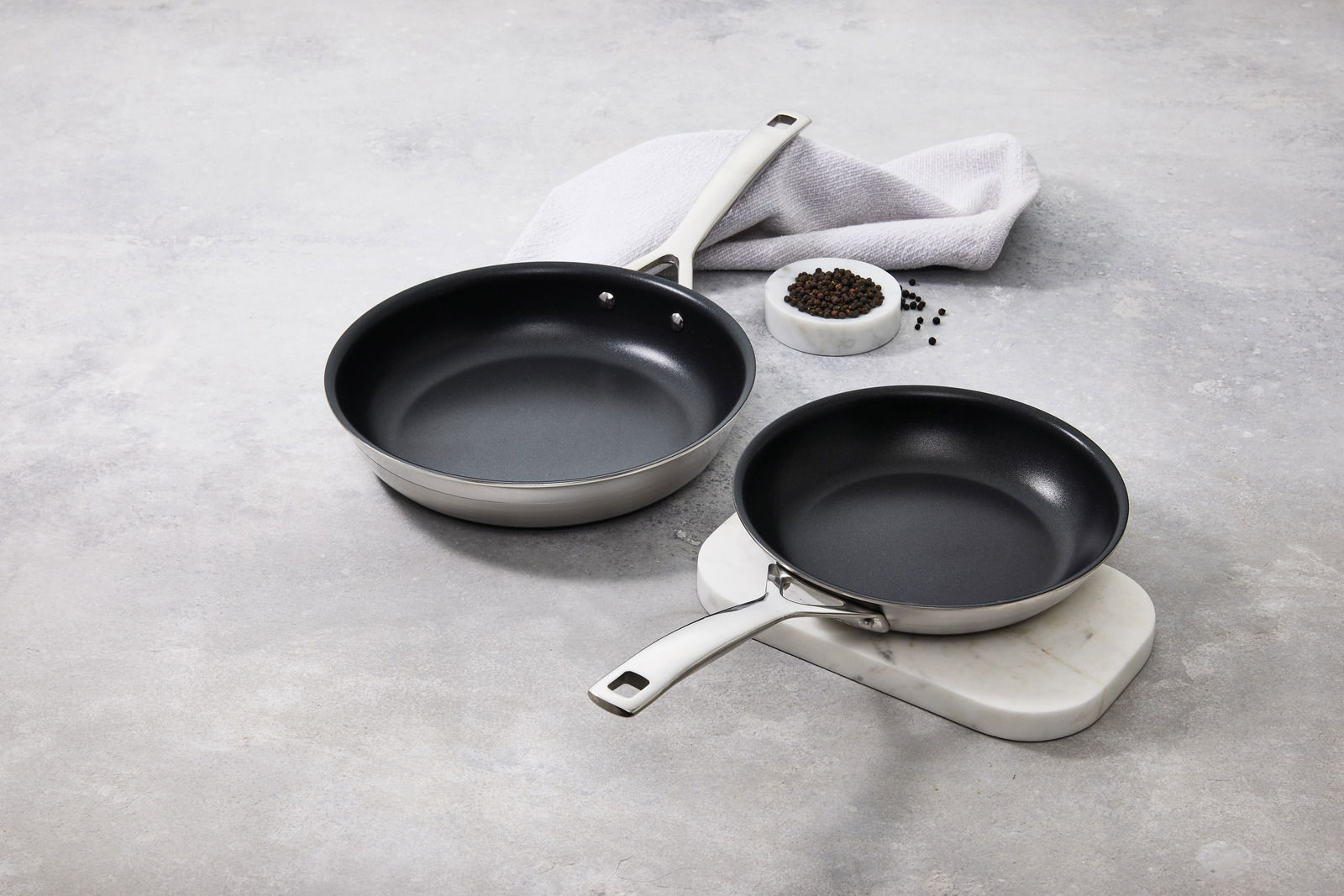 LE CREUSET Pfannenset 2-tlg. 3-PLY Zwei 3-PLY Pfannen in verschiedenen Größen auf einer hellen Oberfläche, mit einer seitlichen Perspektive. Eine Pfanne liegt auf einem Marmorbrett, daneben eine Schale mit Pfefferkörnern und ein weißes Tuch.
