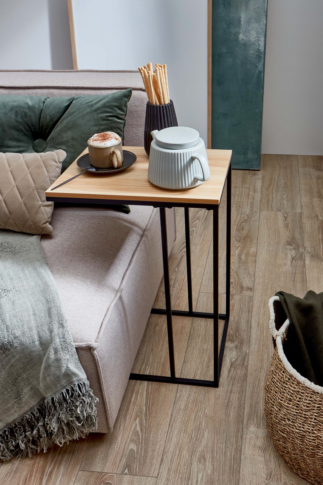 Schmaler Beistelltisch mit heller Holzplatte und schwarzem Metallgestell neben einem Sofa, darauf Tasse und Keramikdose, fotografiert in seitlicher Perspektive auf das Produkt.